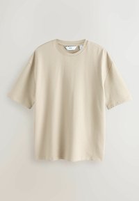 T-shirt beige à manches courtes en maille texturée avec col rond, suspendu devant un fond clair uni.