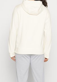 Persona che indossa un felpa con cappuccio color crema e pantaloni della tuta grigio chiaro, mostrata di spalle contro uno sfondo semplice.