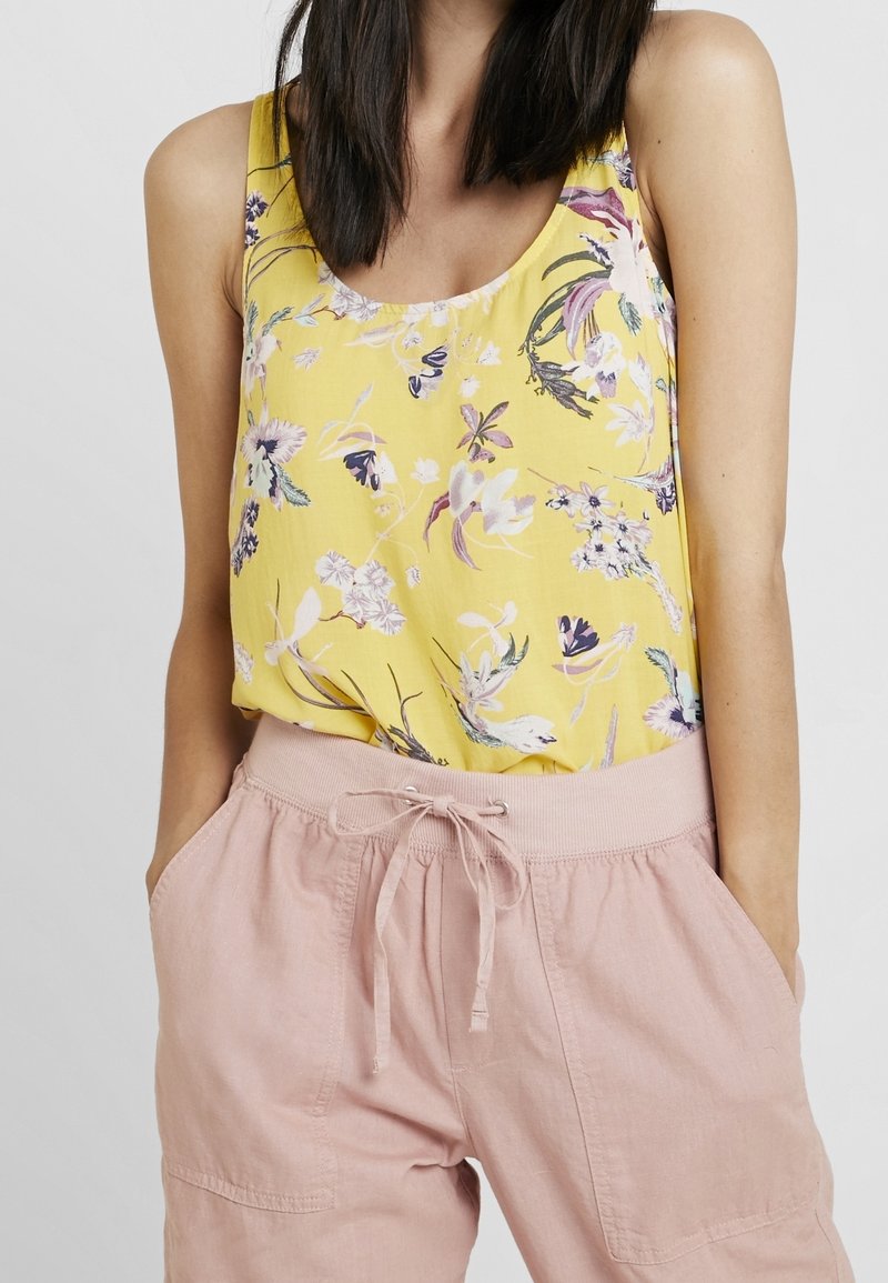 Keltainen kukkakuosiset tanktoppi, valmistettu kevyestä kankaasta, jossa on pyöreä pääntie ja löysä leikkaus. Yhdistettynä vaaleanpunaisiin narusahkoihin shortseihin.