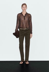Femme portant une chemise longue à manches transparentes marron, un pantalon vert olive, une ceinture cloutée, tenant un chapeau marron, et des sandales noires, se tenant contre un mur blanc.