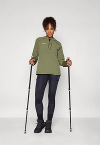 Olive green long-sleeve pullover with quarter zipper, paired with dark leggings and black hiking boots. Equipped with hiking poles.
