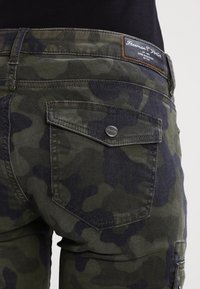 Kamo kalhoty v tmavě zelené a černé barvě, s viditelnou zadní kapsou, kovovými akcenty a koženou nášivkou s logem na pasu.