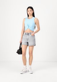 Μπλε αμάνικο crop top με στριμμένο μπροστινό μέρος, ανοιχτό γκρι τζιν σορτσάκι, λευκά ψηλά αθλητικά παπούτσια και μια μαύρη τσάντα με σχέδια.
