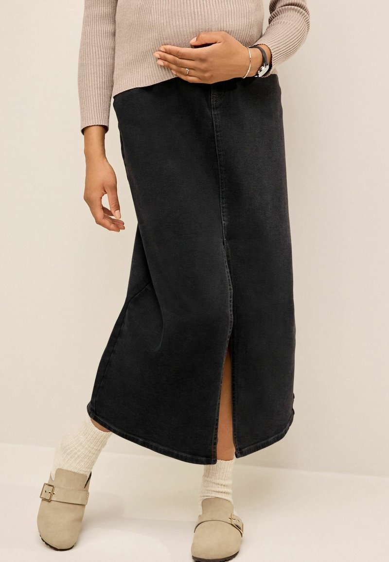 Personne portant un pull beige, une jupe midi en denim noir avec une fente devant, des chaussettes blanches et des chaussures à enfiler beiges avec boucle, se tenant devant un fond uni.