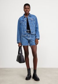 Chaqueta de mezclilla en azul claro, con botones y bolsillos. Combinada con pantalones cortos de mezclilla a juego, top negro y botas negras gruesas. Bolso negro.