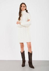 Frau in einem weißen, grob gestrickten Rollkragenkleid und dunkelbraunen, bestickten Cowboy-Stiefeln, die mit gekreuzten Armen dasteht.