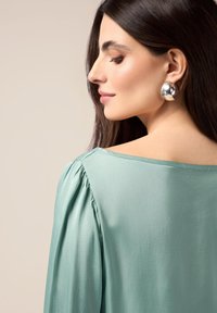Blusa verde menta con una vestibilità rilassata, plissé sulle spalle e scollatura ampia. Orecchini circolari in argento completano il look. Tessuto morbido.