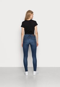 Jeans skinny em denim azul escuro com uma textura suave, combinados com uma blusa de manga curta preta. Bolsos traseiros notáveis e um retalho de couro com marca.