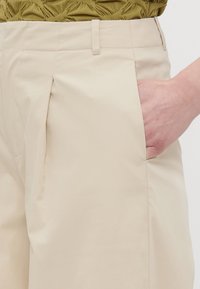 Beige, plisserte bukser med sidelomme og en glatt, lett skinnende tekstur. Midjen har et minimalistisk design med enkel søm.