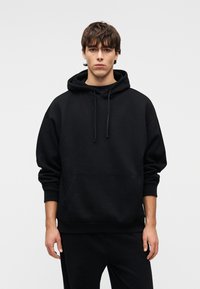 Μαύρο φούτερ με κουκούλα και μεγάλο μπροστινό τσέπη καγκουρό, με κορδόνι στην κουκούλα και ριμπ μανσέτες, κατασκευασμένο από απαλό, υφασμάτινο ύφασμα με υφή.