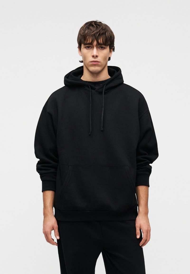 Μαύρο φούτερ με κουκούλα και μεγάλο μπροστινό τσέπη καγκουρό, με κορδόνι στην κουκούλα και ριμπ μανσέτες, κατασκευασμένο από απαλό, υφασμάτινο ύφασμα με υφή.