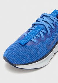 Blue athletic shoe featuring a textured knit upper, tonal laces, and a cushioned sole with a contrasting black accent along the edge.