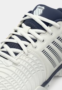 Încălțăminte sport cu un design alb și albastru închis, având un material superior texturat, șireturi contrastante și un logo în formă de scut pe limbă.