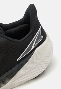 Chaussure de sport noire avec un dessus en tissu texturé, un col rembourré et une semelle en caoutchouc blanc avec des rainures diagonales. Détails du logo en argent.