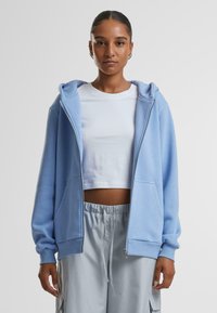 Felpa azzurra con zip e cappuccio, polsini a coste e tasche frontali, indossata sopra una maglietta bianca corta e pantaloni grigi chiari.