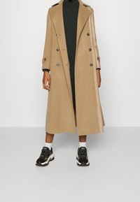 Manteau trench beige long en mélange de laine, avec un design double boutonnage, de larges revers et des boutons décoratifs ; associé à des baskets noires.