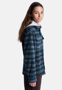 Fleecejacka med ett teal- och lila geometriskt mönster, knäppning fram, kontrasterande grå huva och fickor fram.