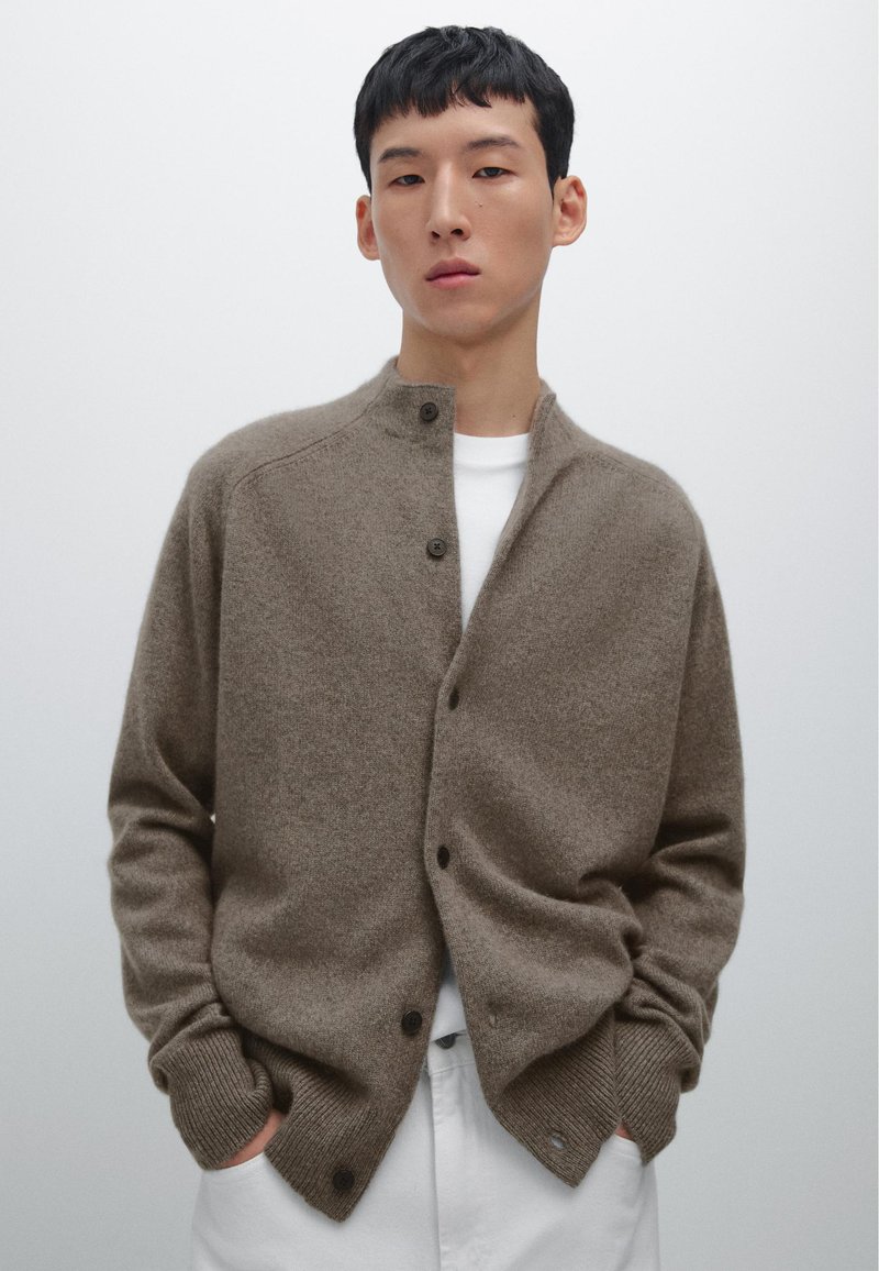 Cardigan marrone, texturizzato, con colletto alto, caratterizzato da bottoni scuri lungo la parte anteriore e polsini a coste. Indossato su una camicia bianca.