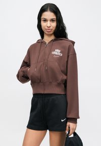 Νεαρή γυναίκα που φοράει καφέ φούτερ με φερμουάρ Nike Athletics και μαύρο σορτς Nike, κρατώντας ένα μαύρο καπέλο στο δεξί της χέρι.