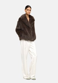 Brązowy płaszcz z futra syntetycznego z dużym kołnierzem, zestawiony z białymi szerokimi spodniami i miękkimi zielonymi butami. Płaszcz ma fakturowane wykończenie.