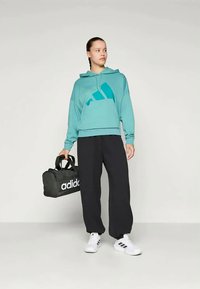 Teal cropped hoodie with a large logo, black loose-fit pants, white sneakers, and black duffle bag with white branding.