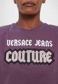 Lilla bomull t-skjorte med hvit og svart brodert tekst "VERSACE JEANS COUTURE" med rhinestones på en svart lapp.