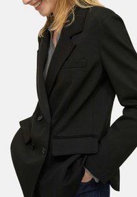 Blazer nero su misura con design doppiopetto, due tasche frontali e rever strutturati, con una texture del tessuto liscia.