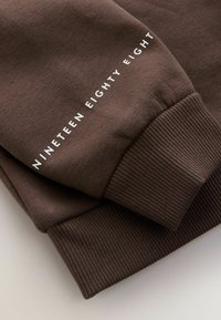 Hnědý svetr s ribbovaným manžetem; má bílý text na délku "NINETEEN EIGHTY EIGHT". Hladká textilie.