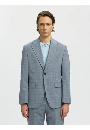 Mann trägt hellgrauen karierten Anzug und hellblaues Zip-up-Hemd, steht vor schlichtem weißem Hintergrund.