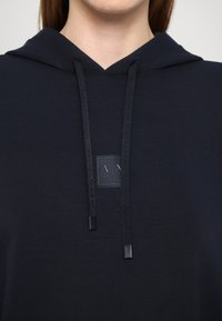 Mustapaita, jossa on nyöri, ja rintataskussa on teksturoitu logopatch. Silkkinen kangas, minimalistinen muotoilu ja istuva siluetti.