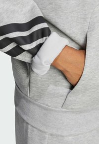 Sivi dukserica s crnim trakama na rukavima, s rebrastim manžetama i džepom. Materijal izgleda mekano, s udobnom teksturom.