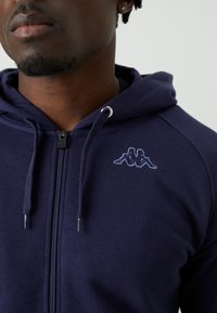 Námornícke zip-up mikina z mäkkého materiálu s kloktaním vreckom, s malým Svetlomodrým vyšitým logom na ľavej hrudi a strieborným zipsom.