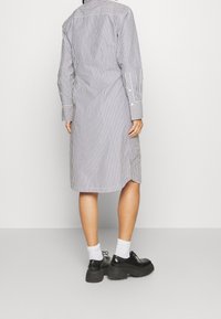 Persona che indossa un abito a righe verticali, lungo fino al ginocchio, con maniche lunghe, calze bianche e scarpe chunky nere, in piedi contro uno sfondo neutro.