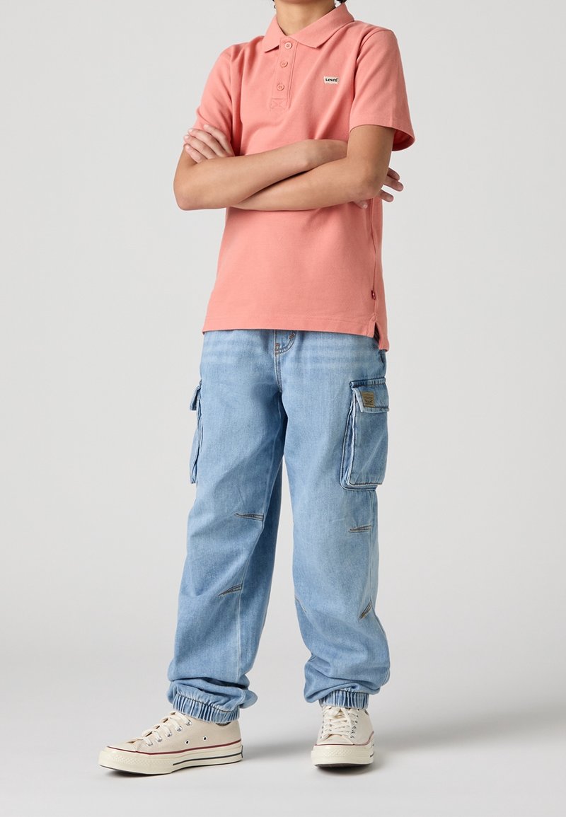 Personne portant un polo rose corail et un jean cargo bleu clair avec des poignets élastiques, debout les bras croisés, baskets blanches visibles.
