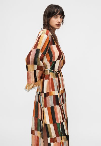 Γυναίκα με σκούρα μαλλιά που φοράει πολύχρωμο φόρεμα πατςγουορκ με φαρδιά μανίκια και κρόσσια, που στέκεται πλαγίως μπροστά σε ένα απλό φόντο.