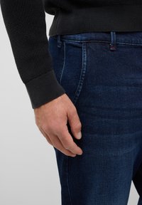 Dunkelblaue Jeans aus Denim mit glatter Textur und dezentem Verblassen, getragen mit einem schwarzen Strickpullover mit gerippten Bündchen.
