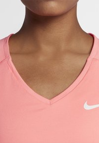 Top deportivo de color rosa con escote en V, hecho de una tela suave. Presenta mangas cortas y un logo blanco de Nike en el lado izquierdo. Textura lisa.