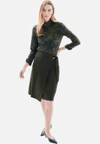 Haut à manches longues vert à motifs associé à une jupe portefeuille olive foncé avec une boucle avant et un long lien, portée avec des talons noirs.