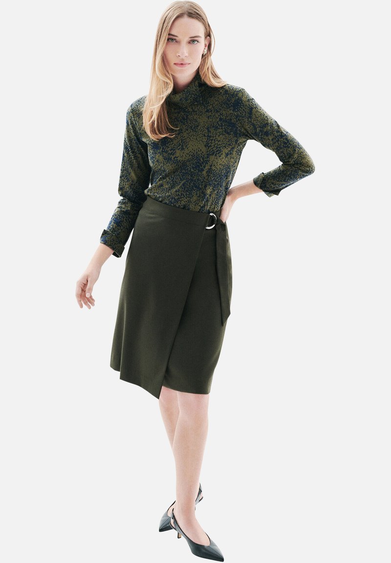 Haut à manches longues vert à motifs associé à une jupe portefeuille olive foncé avec une boucle avant et un long lien, portée avec des talons noirs.