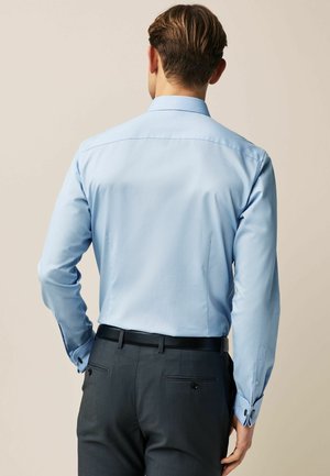 Himmelblaues Hemd mit langen Ärmeln, klassischem Kragen und kontrastierenden dunklen Knöpfen, kombiniert mit grauen Anzughosen und einem schwarzen Gürtel.