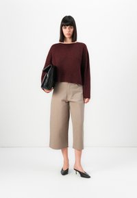 Femme aux cheveux noirs raides portant un pull bordeaux, un pantalon beige court, des talons noirs et tenant un grand sac à main noir.