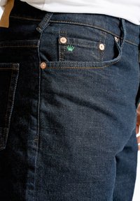 Dunkelblaue Denim-Jeans mit Kupferriegeln, Kontrastnähten und einem kleinen grünen Logo auf der Gesäßtasche. Schmaler Schnitt mit detaillierter Textur.