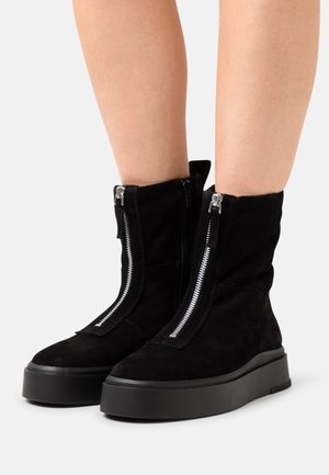 Bottines en daim noir avec deux fermetures éclair argentées, une semelle plateforme noire épaisse et un design légèrement décontracté. Détail du matériau texturé visible.