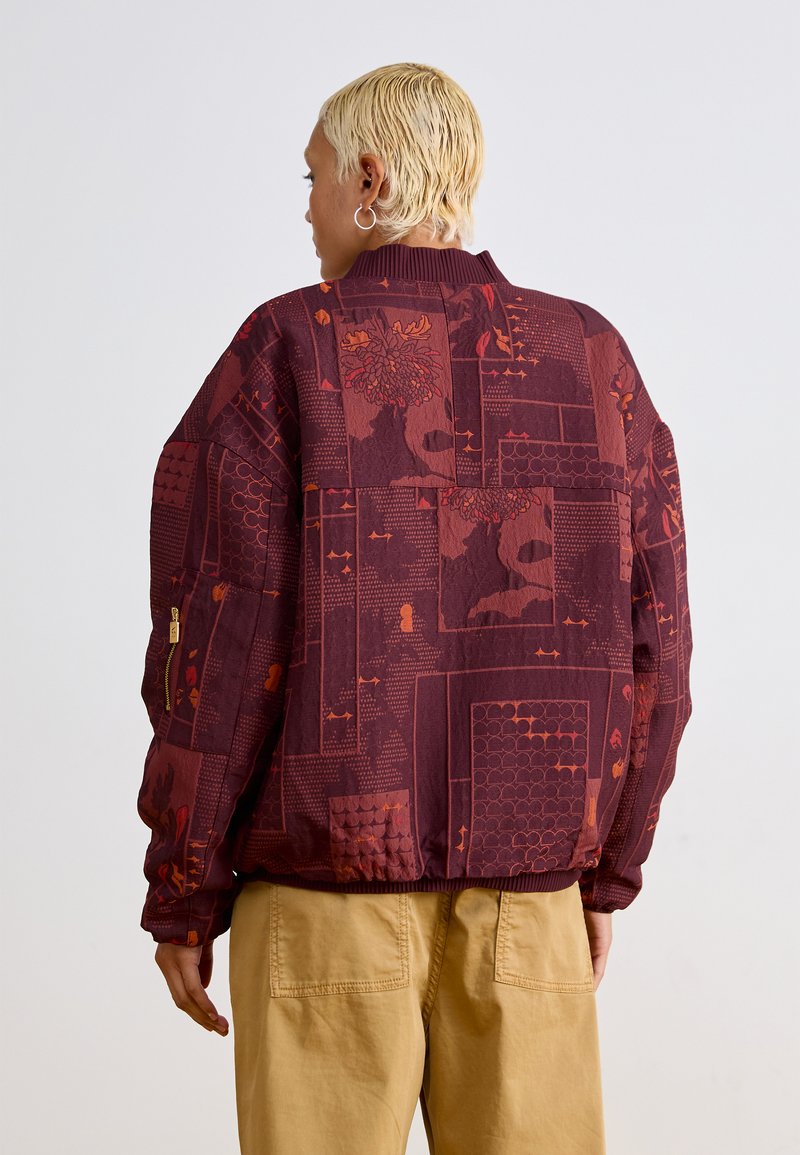 Bordeauxfarbige Jacke aus strukturiertem Stoff mit verschiedenen Mustern in roten und orangefarbenen Tönen. Gerippte Bündchen und Saum, seitliche Reißverschlusstaschen.