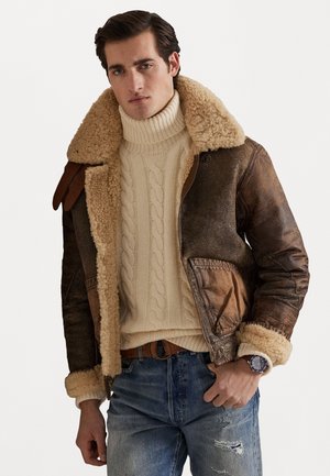 Giacca bomber in pelle marrone con colletto e polsini in shearling crema, indossata sopra un maglione a collo alto in maglia cable-knit crema, abbinata a jeans blu.