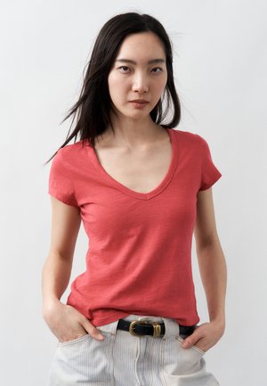 Mlada ženska z dolgimi temnimi lasmi nosi koralno T-majico z V-izrezom, speto v svetle hlače z črtami in črnim pasom, stoji z rokami v žepih.