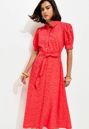 Kobieta w czerwonej sukience z paskiem, z bufiastymi rękawami i kwiatowym wzorem ażurowym, ozdobiona złotymi i beżowymi bransoletkami.