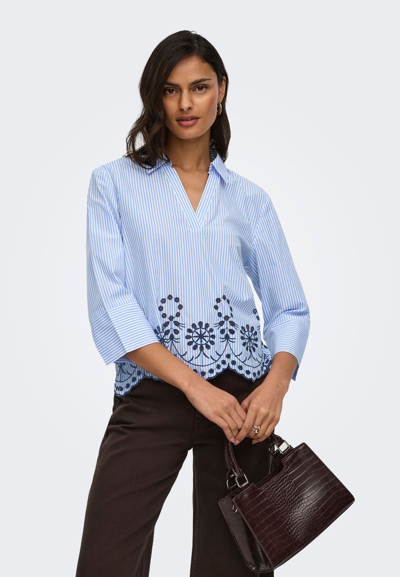Femme portant un chemisier rayé bleu et blanc avec broderie florale et un pantalon noir, tenant un sac à main marron foncé avec deux poignées.