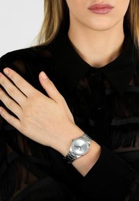 Silverklocka med en texturerad rund urtavla och kristalldetaljer, monterad på ett armband av metall länkar, bärs på en hand mot en svart blus.