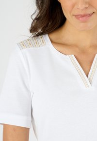 Witte katoenen T-shirt met een V-hals en gouden en beige gestreepte accenten op de schouders en opgerolde mouwen, zachte textuur.
